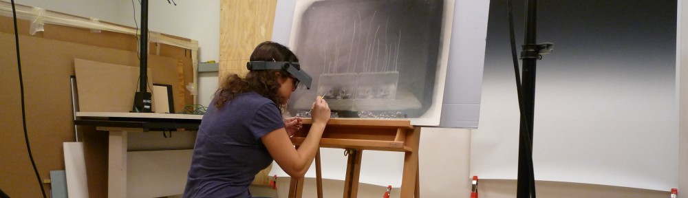 Image of Lisa Duncan wearing a headband magnifier and working at an easel. She is performing a conservation treatment on a large black and white artwork.
